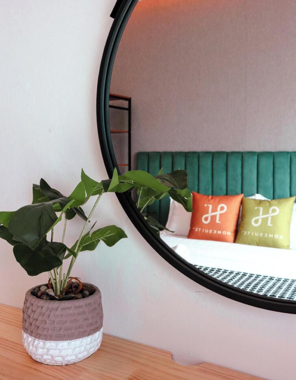 A bedroom with a green upholstered headboard, a round mirror, and a potted plant on a wooden nightstand.