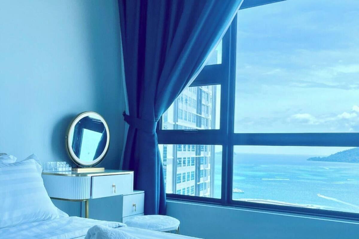 Une chambre d’hôtel avec un lit, une table de chevet avec un miroir, une fenêtre avec des rideaux et une vue sur la mer et les bâtiments.