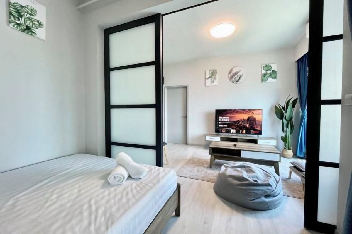 A modern hotel room with a bed, a television, a bean bag, and a door with a black frame.