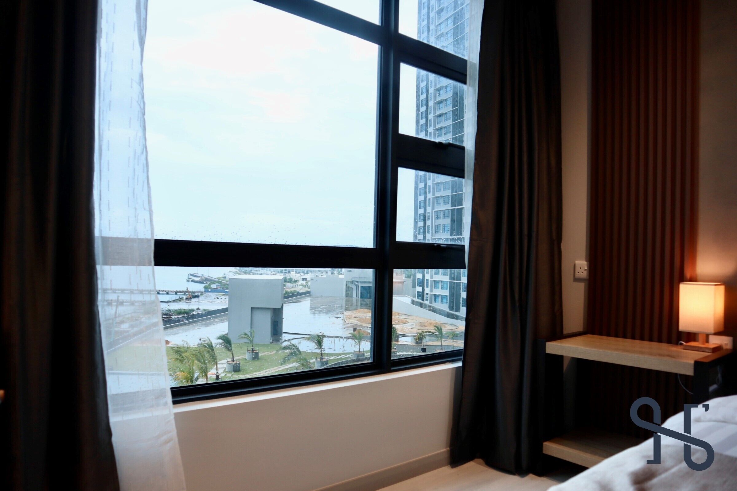 Ein Hotelzimmer mit großem Fenster, Nachttisch mit Lampe und Blick auf eine Stadtlandschaft.