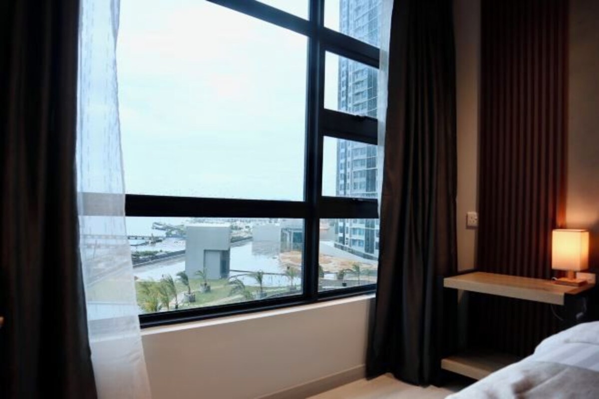 Quarto de hotel com uma janela grande, uma mesa de cabeceira com abajur e vista para a cidade.
