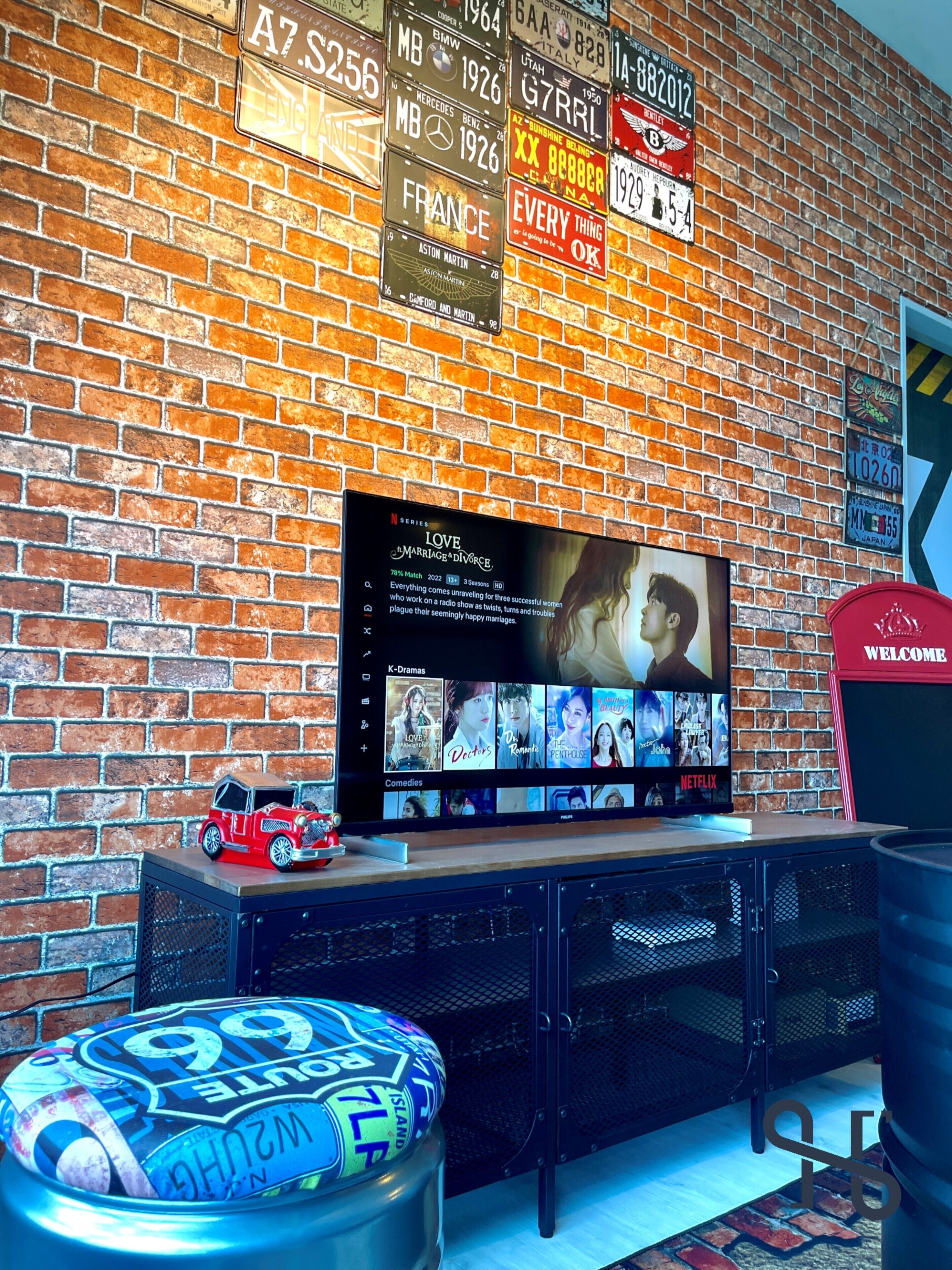 A brick wall with vintage license plates, a flat-screen TV displaying a Netflix show, and a small red toy car on a shelf.