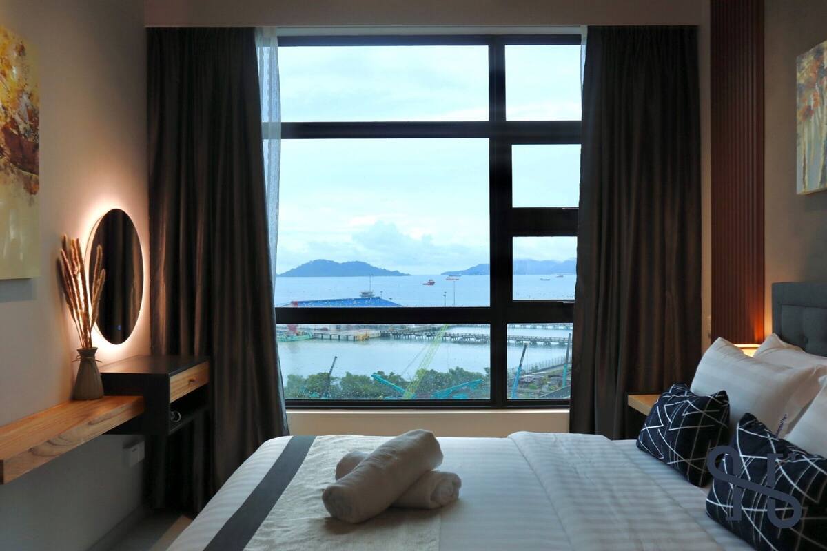 Une chambre d’hôtel dotée d’une grande fenêtre, d’un lit avec des oreillers, d’une table de chevet avec une lampe, et offrant une vue sur la mer et un pont.