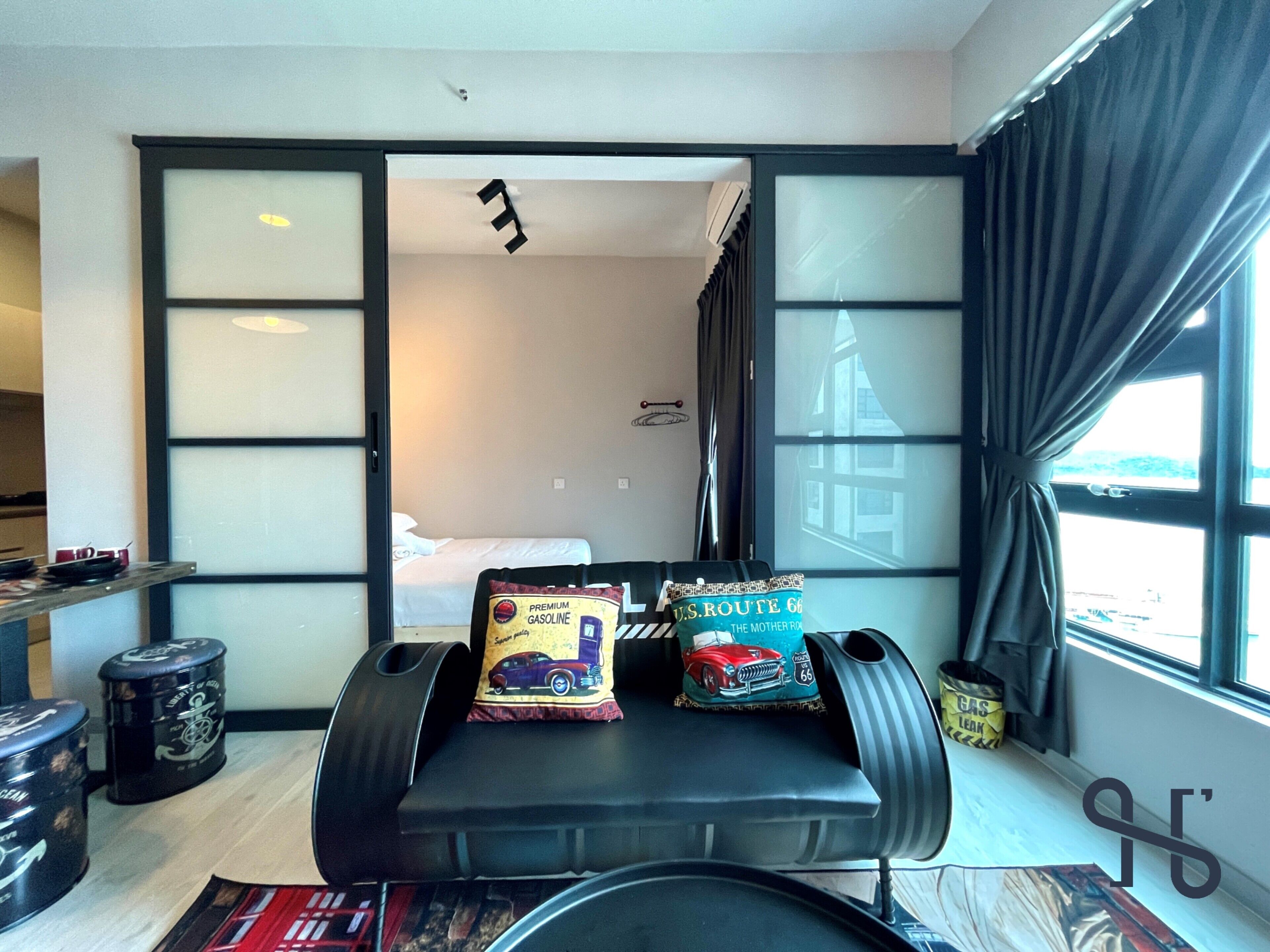 A compact hotel room with a bed, a small table with snacks, a trash bin, and a window with curtains.