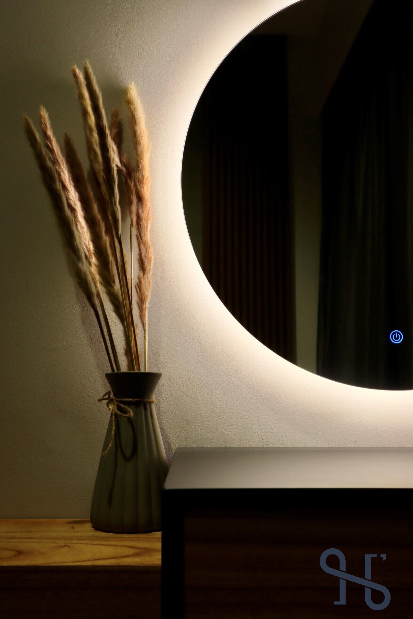 A vase with pampas grass on a wooden surface next to a wall with a circular mirror.