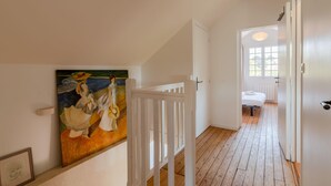 A hallway with a white railing, a painting on the wall, and a view into a bedroom with a bed and a window. - La Paisible - 50m mer - Jardin (La Bernerie-en-Retz)