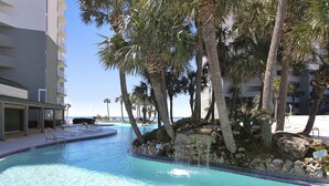 A swimming pool with a waterfall feature, surrounded by palm trees and a building with balconies.