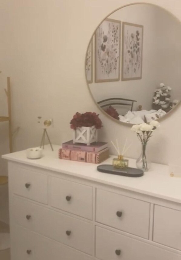 Et hvidt kommode med et rundt spejl, en lille hvid vase med blomster og nogle få dekorative genstande ovenpå.