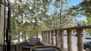 A balcony with wicker furniture and a glass table, overlooking a street with cars and trees.