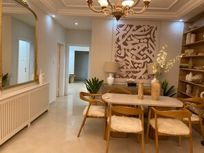 A dining area with a white table and chairs, a chandelier, a large abstract wall art, and a bookshelf with decorative items. - Sleeps 8 people! Apartment 5 minutes to Abdoun w balcony a in Dair Ghbar!  (Amman)