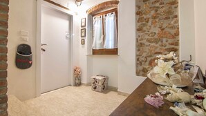 Una habitación con puerta blanca, pared de piedra y techo de madera. Hay una mesita con un jarrón de flores y una caja decorativa.