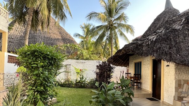 A thatched-roof structure with a tiled entrance and a small garden area with palm trees and various plants. - 1 Bed Villa - Sleeps 2 - Pool - Smoking - Parking (Diani Beach kenya)