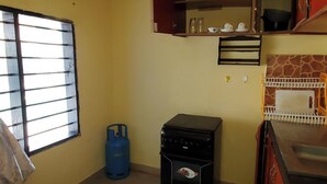 A compact kitchen with a gas stove, a blue gas cylinder, a microwave, and a window with bars. - 1 Bed Villa - Sleeps 2 - Pool - Smoking - Parking (Diani Beach kenya)