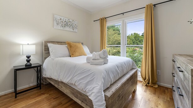 Een slaapkamer met een houten bedframe, witte beddengoed, een nachtkastje met een lamp, en een raam met gordijnen.