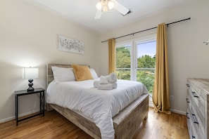 A bedroom with a wooden bed frame, white bedding, a bedside table with a lamp, and a window with curtains.
