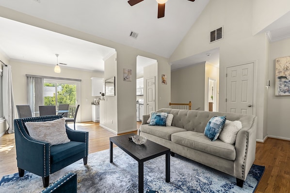 A spacious living room with a sofa, armchair, and coffee table. There is a dining area with chairs and a table in the background.