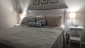 Ein ordentlich bezogenes Bett mit einer gesteppten Tagesdecke, mehreren Kissen, einer Nachttischlampe, einem kleinen Nachttisch und drei gerahmten Bildern an der Wand.