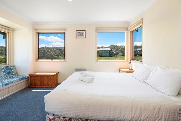 A bedroom with a large bed, a wooden bench, and two windows offering views of the outdoors.