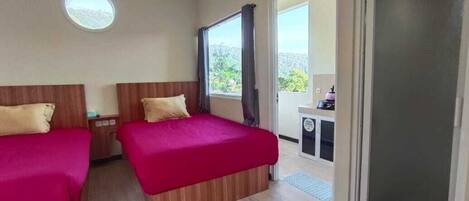 A room with two beds, a small desk, a round window, and a balcony with a view.
