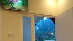 A corridor with a television mounted on the wall, a door leading to a balcony with a red welcome mat, and a view of a garden with a red-roofed structure.