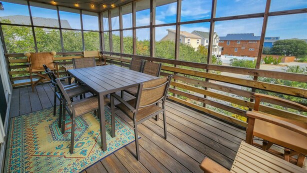 Eine überdachte Terrasse mit Holztisch und -stühlen, einem gemusterten Teppich und Blick auf Häuser im Hintergrund.