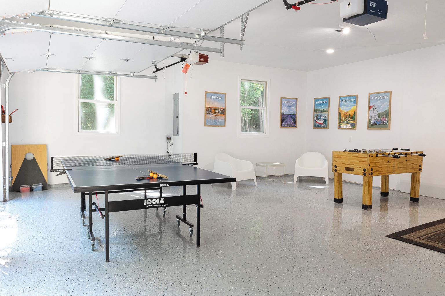 A room with a ping pong table, two white chairs, a foosball table, and a projector mounted on the ceiling.