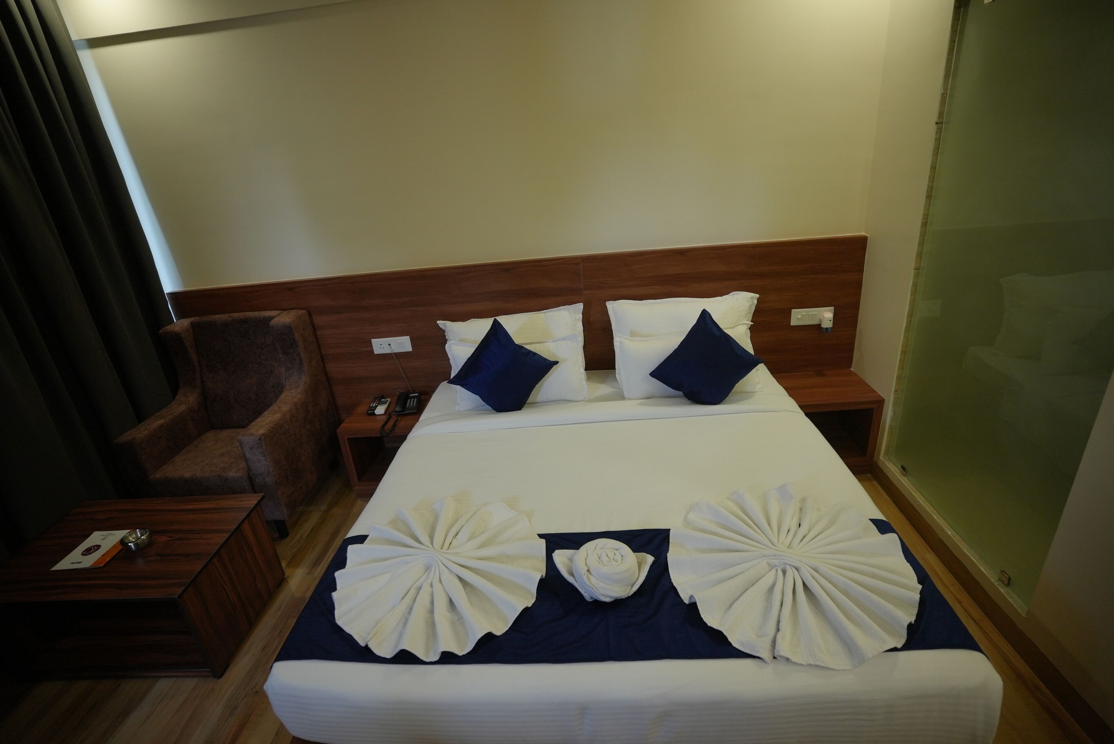 Une chambre d’hôtel avec un grand lit, une tête de lit en bois, un fauteuil marron, une table de chevet en bois et une cloison en verre.