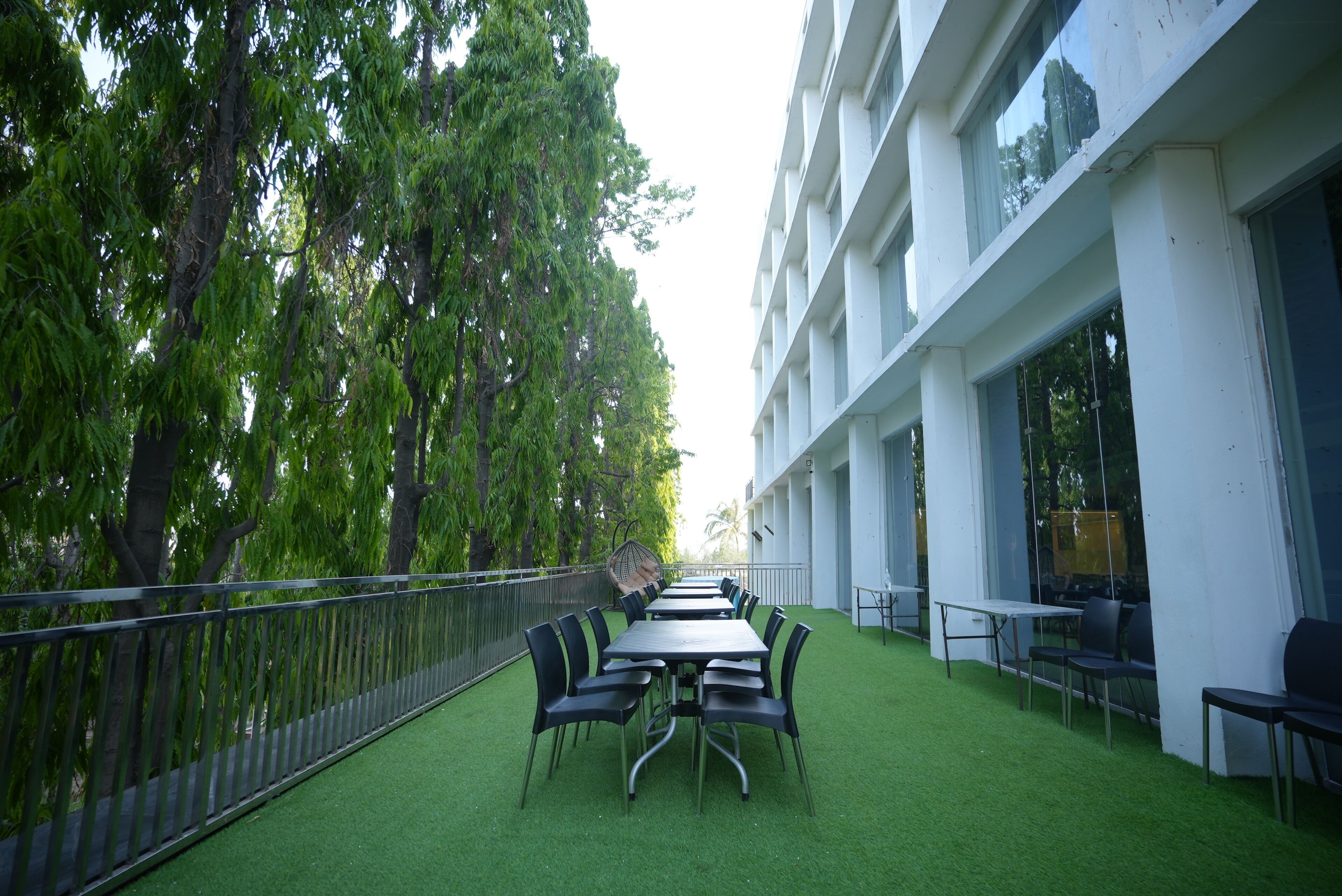 An outdoor terrace with a long table and chairs, surrounded by lush greenery and a white building.