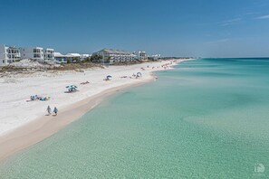 A beach with clear turquoise water, white sand, and beach umbrellas. There are buildings with balconies along the shore.