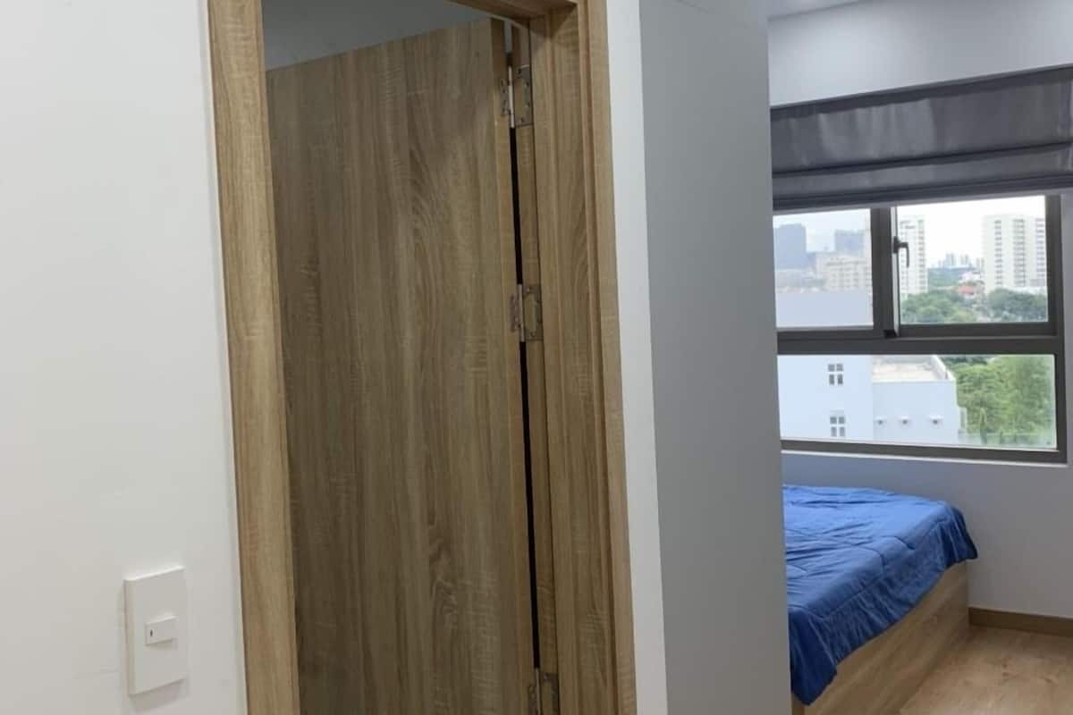 A room with a wooden wardrobe, a bed with a blue bedspread, a window with a view of buildings, and a light switch on the wall.