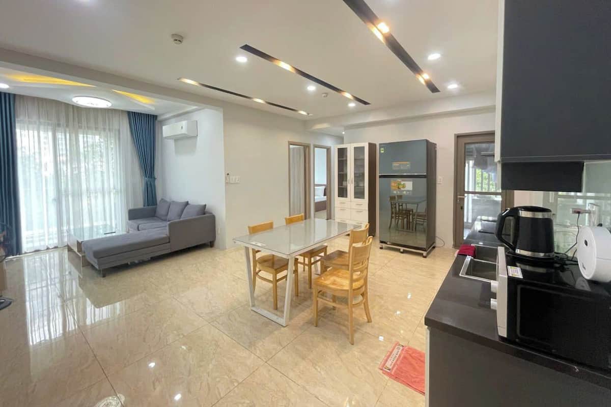 A modern living room with a glass dining table, a sofa, and a kitchen area with a coffee machine.