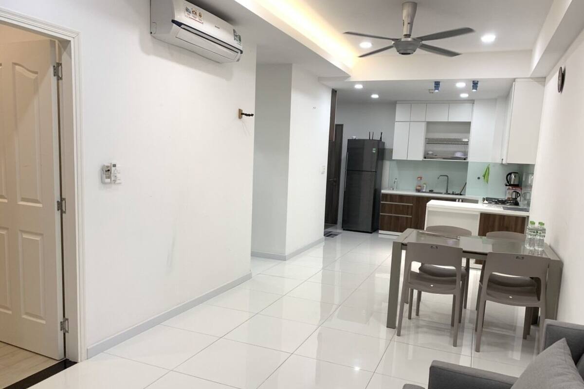 A modern living room with a sofa and a dining table with chairs. There is a kitchen area with cabinets and a refrigerator.