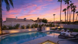 A swimming pool with lounge chairs and a hot tub, surrounded by palm trees and buildings.