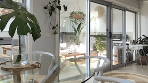 A modern living room with a glass dining table, white chairs, and a large potted plant.