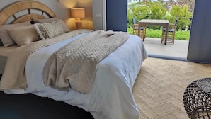 A bedroom with a large bed, a bedside lamp, and a view of an outdoor patio with a table and chairs.