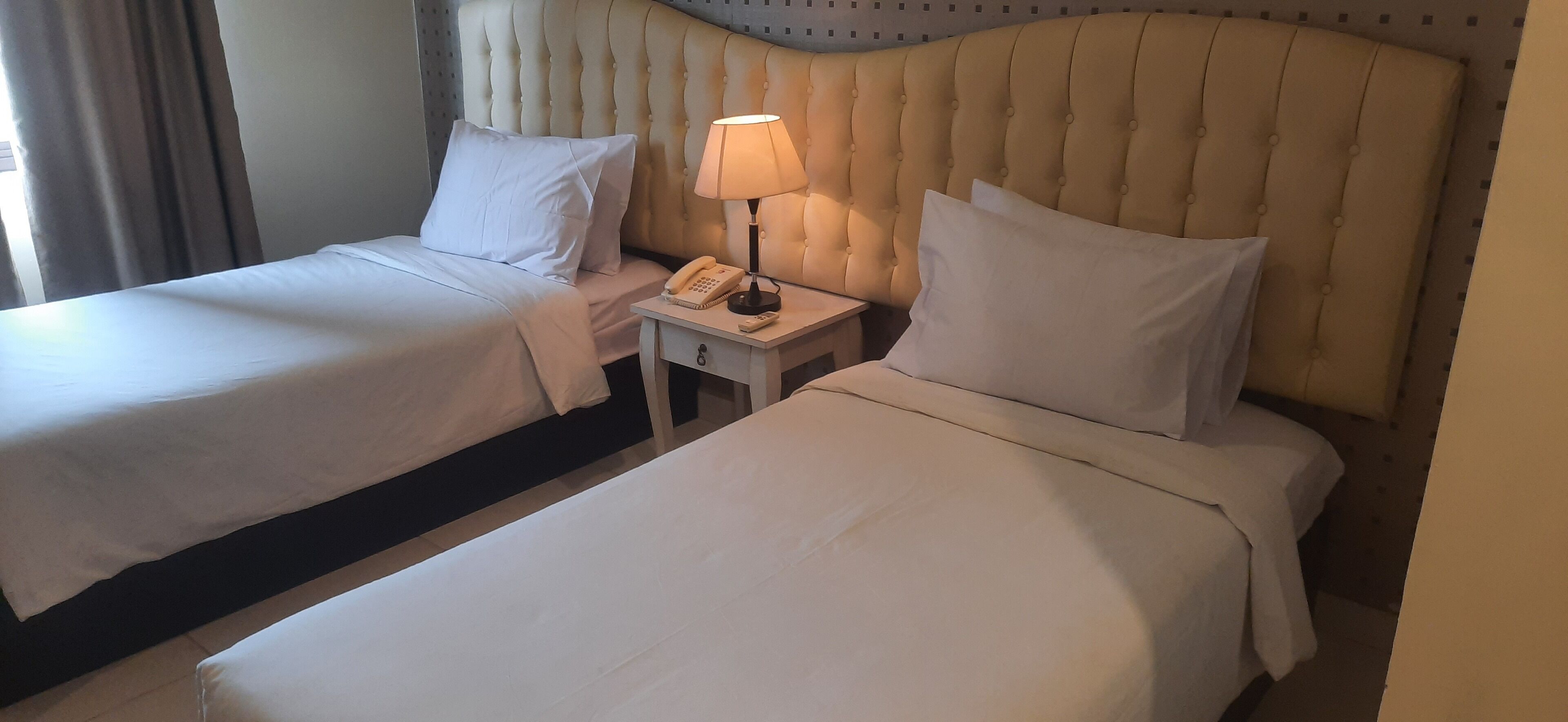 A hotel room with two beds, each with white linens and pillows, a bedside table with a lamp and a phone, and a tufted headboard.