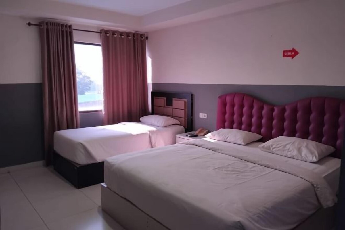 Hotelkamer met twee bedden, een groot raam met gordijnen, een aan de muur bevestigd telefoon en een rood bord dat de richting naar de badkamer aangeeft.