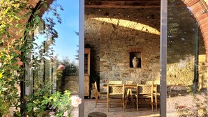 An outdoor patio area with a stone wall, wooden furniture, and a glass door leading to a room. - Exclusive Tuscan Winery Escape: Charming Villa with Pool & Breathtaking Views. (Radda in Chianti)