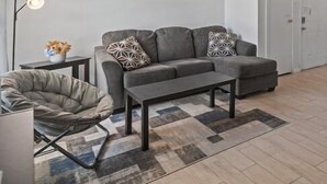 A modern living room with a grey sofa, a coffee table, and a unique chair. A painting of trees hangs on the wall, and a floor lamp is visible. - Cozy 2 Bed 1 Bath Home near IAH (Spring)