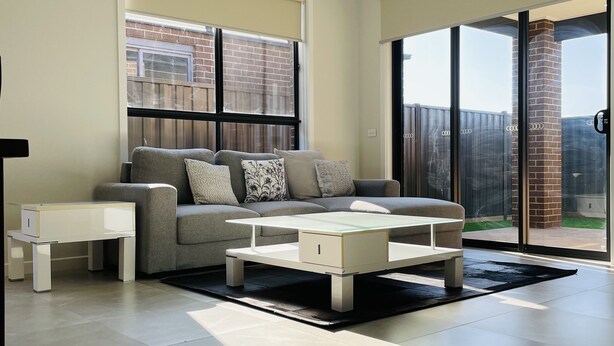 Ein modernes Wohnzimmer mit grauem Sofa, weißem Couchtisch und einem Tisch mit Glasplatte. Ein großes Schiebeglastür führt auf eine Terrasse.