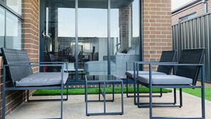 Eine Terrass mit Gartenmöbeln und einer Glastür, die in einen Wohnbereich führt, mit einer Couch und zwei Sesseln.