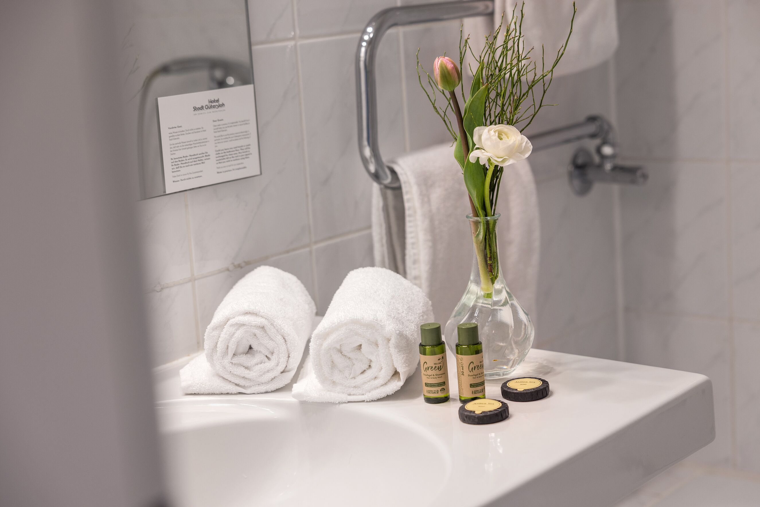 Ein Badezimmer mit einem weißen Waschbecken, zwei aufgerollten Handtüchern, einer Vase mit Blumen und zwei Lotionflaschen.