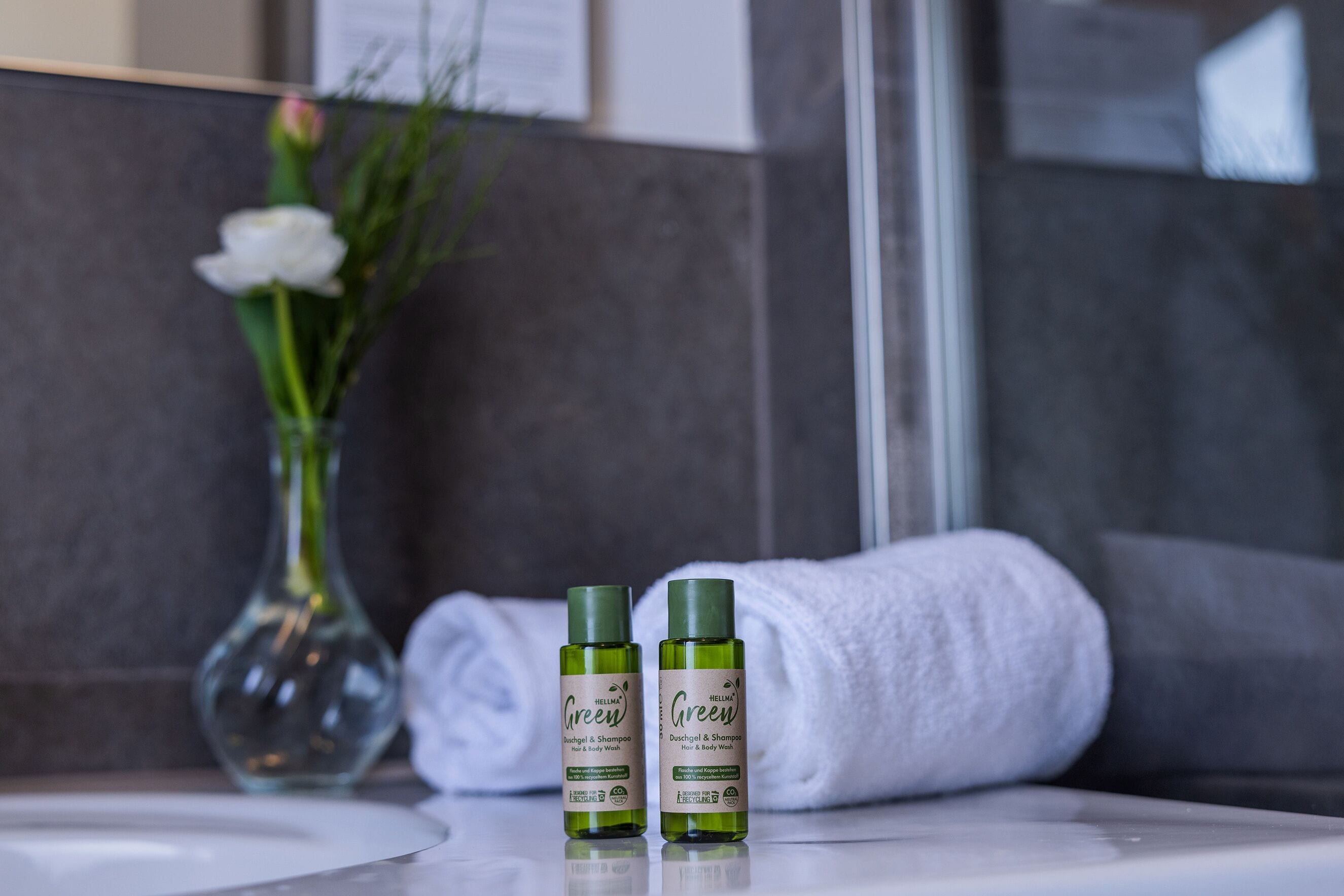 Twee flessen 'Green' haar- en bodywash op een badkamermeubel, met een witte handdoek en een bloemenvaas op de achtergrond.