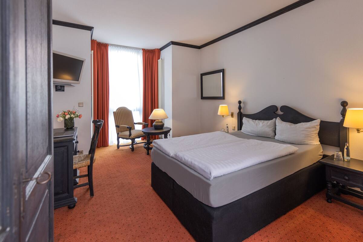 Habitación de hotel con una cama grande, un escritorio con lámpara, una silla, un televisor y una ventana con cortinas.