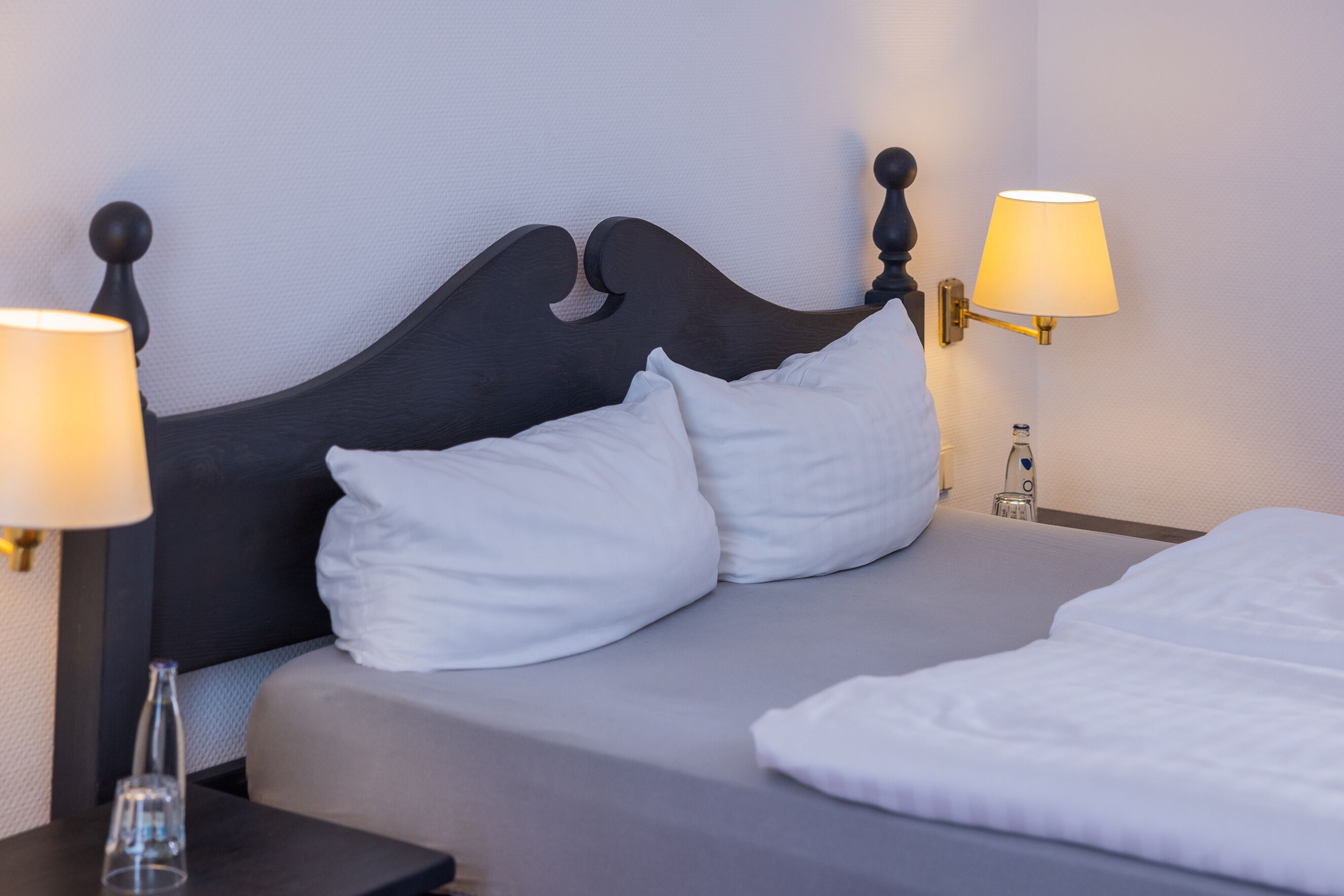 A neatly made bed with white bedding and two pillows, a bedside table with a lamp, and a wall-mounted lamp.
