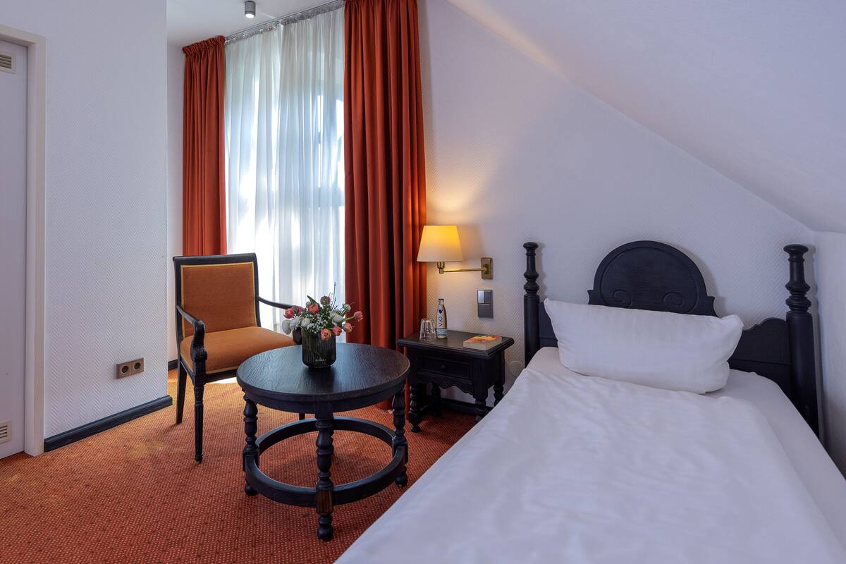 Habitación de hotel con cama, una silla, una mesita con un jarrón de flores, una mesita de noche con una lámpara y una ventana con cortinas.