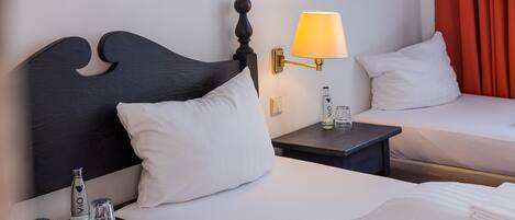 A hotel room with two beds, a bedside table with a bottle and glasses, a lamp, and a wall-mounted mirror.