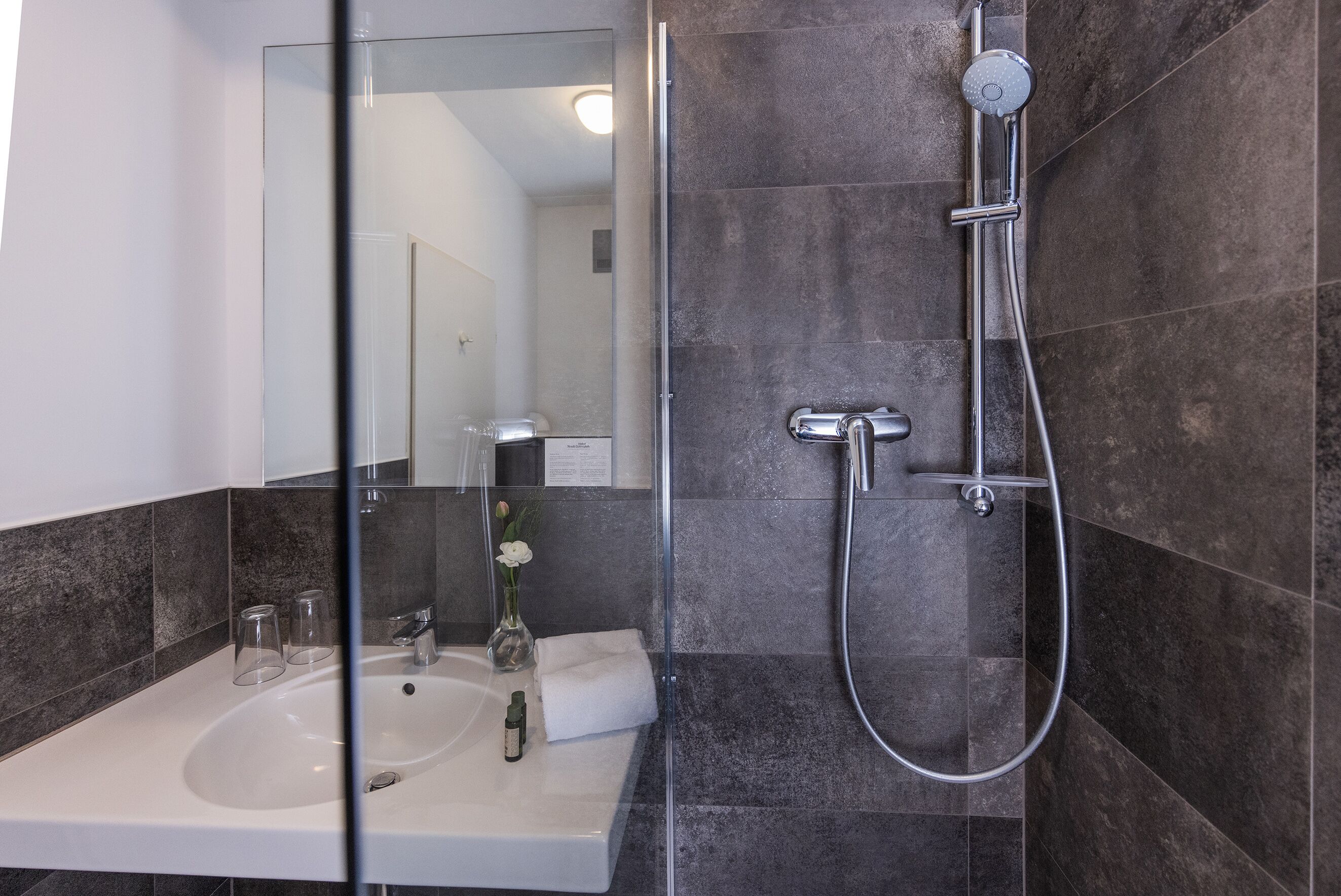 A modern bathroom with a glass shower enclosure, a white sink, and dark tiled walls.