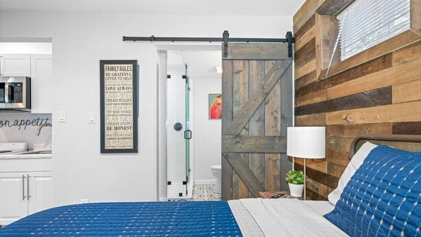 Ein Schlafzimmer mit einem Bett, einer Holzvertäfelung, einer Schiebetür, einer weißen Lampe und einem durch eine offene Tür einsehbaren Küchenbereich.