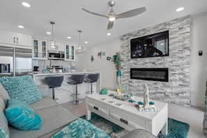 A modern kitchen with white cabinets, a dining area with bar stools, a white console table with decorative items, and a mounted flat-screen TV on a stone-textured wall.